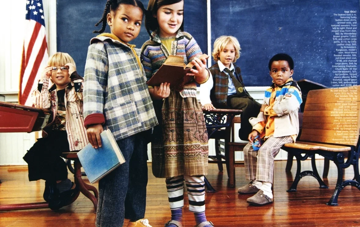 Children in a classroom, with some seated and some standing, near a chalkboard, and an American flag in the corner.