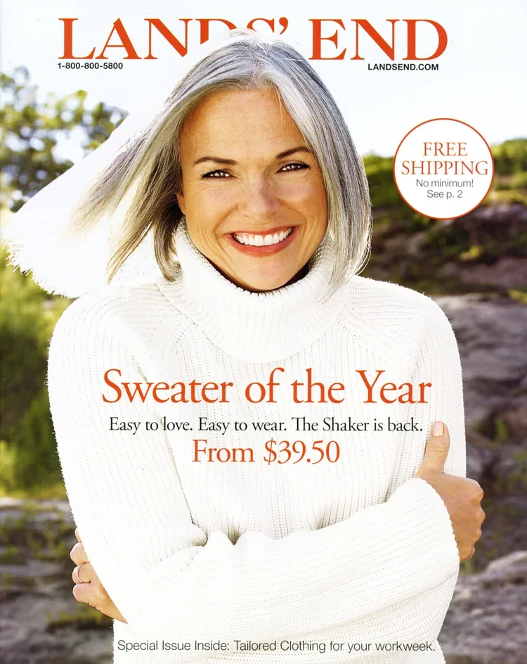 A woman with shoulder-length gray hair smiling and wearing a white turtleneck sweater outdoors with a rocky and green background.