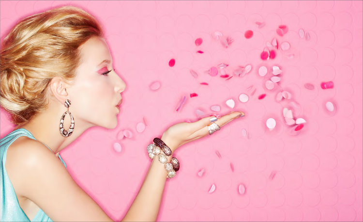 Side view of a woman with styled blonde hair, wearing large earrings and bracelets, blowing pink and white confetti against a pink background.