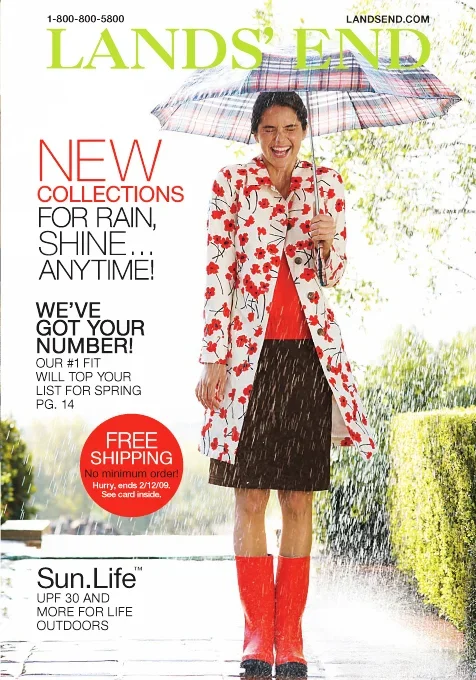 Woman standing in rain holding an umbrella, wearing a floral raincoat, red top, black skirt, red rain boots, smiling outdoors.