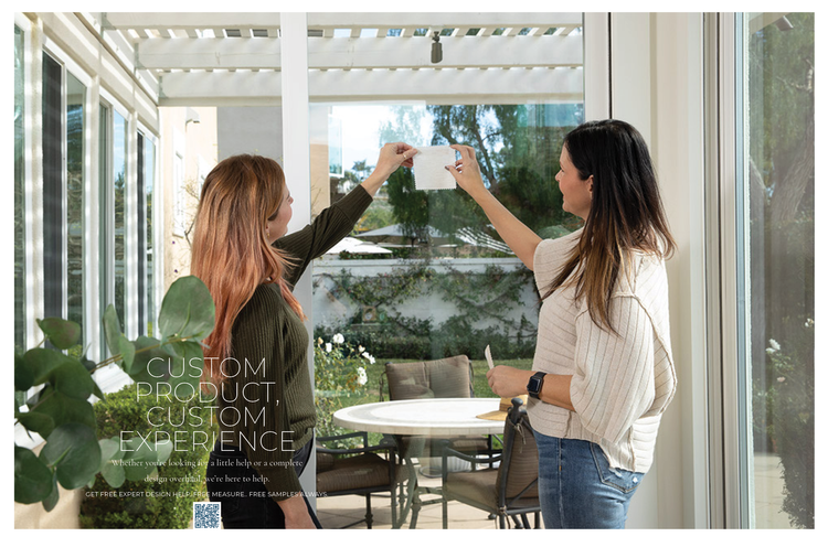 Two women are standing inside near large glass windows, one holding a small square tile while the other points at it, possibly discussing or demonstrating a design or decor idea.