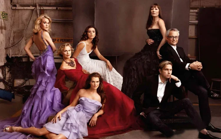 Seven elegantly dressed people, five women and two men, pose together in a professional photoshoot setting. The women wear striking gowns in purple, white, red, and black, while the men are in suits.