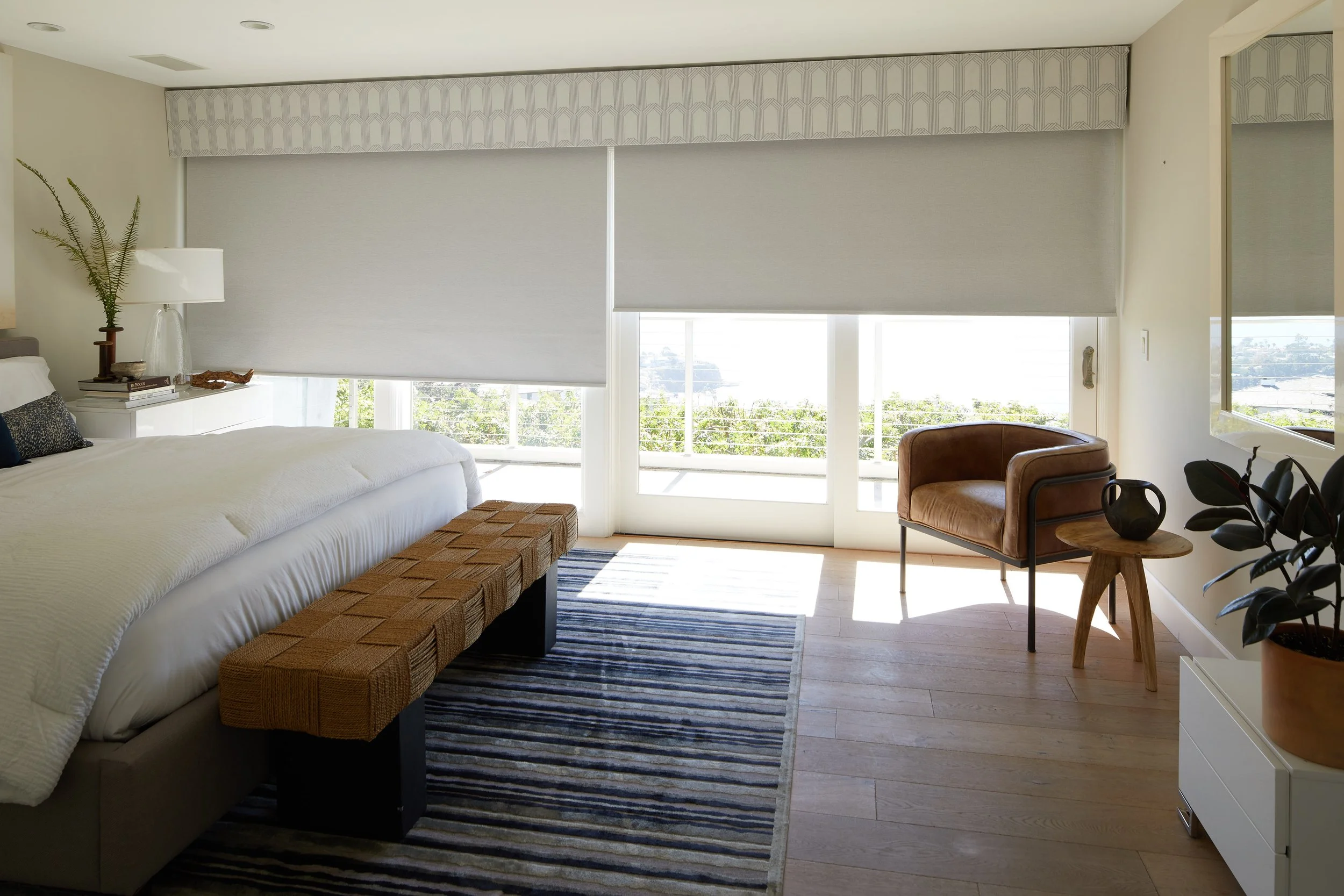 A bedroom with large windows covered by roller blinds. The room has a bed, a wicker bench, a striped rug, a side table with a lamp, a leather chair, and decorative items including plants and vases.