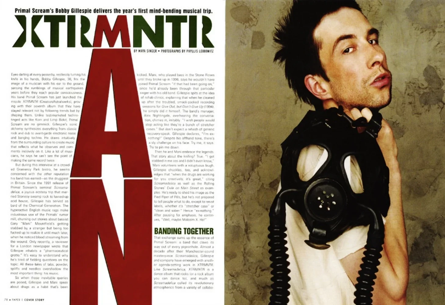 Magazine page featuring Bobby Gillespie from Primal Scream with dark spiky hair, wearing a white shirt and a gas mask hanging around his neck, against a concrete wall background.