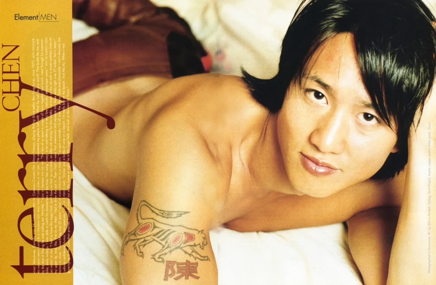 Close-up of a young Asian man with black hair lying on a bed, smiling slightly, with a tattoo of a tiger and Chinese characters on his upper arm.