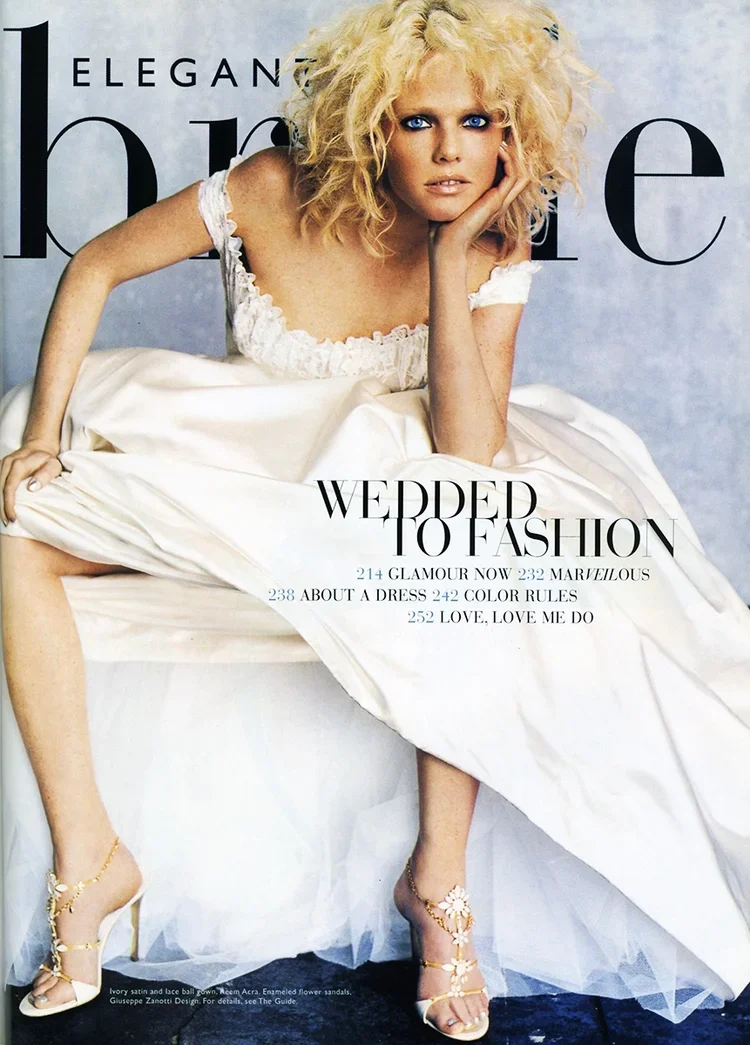 Cover of a fashion magazine featuring a woman with blonde curly hair sitting on a white bed, wearing a white gown and floral high heels, with text about fashion topics.