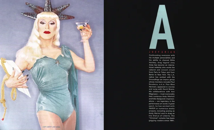 Drag queen with long blonde hair, makeup, a star-shaped headpiece, wearing a teal one-piece costume, holding a cocktail in one hand and a clutch in the other, standing against a gray background.