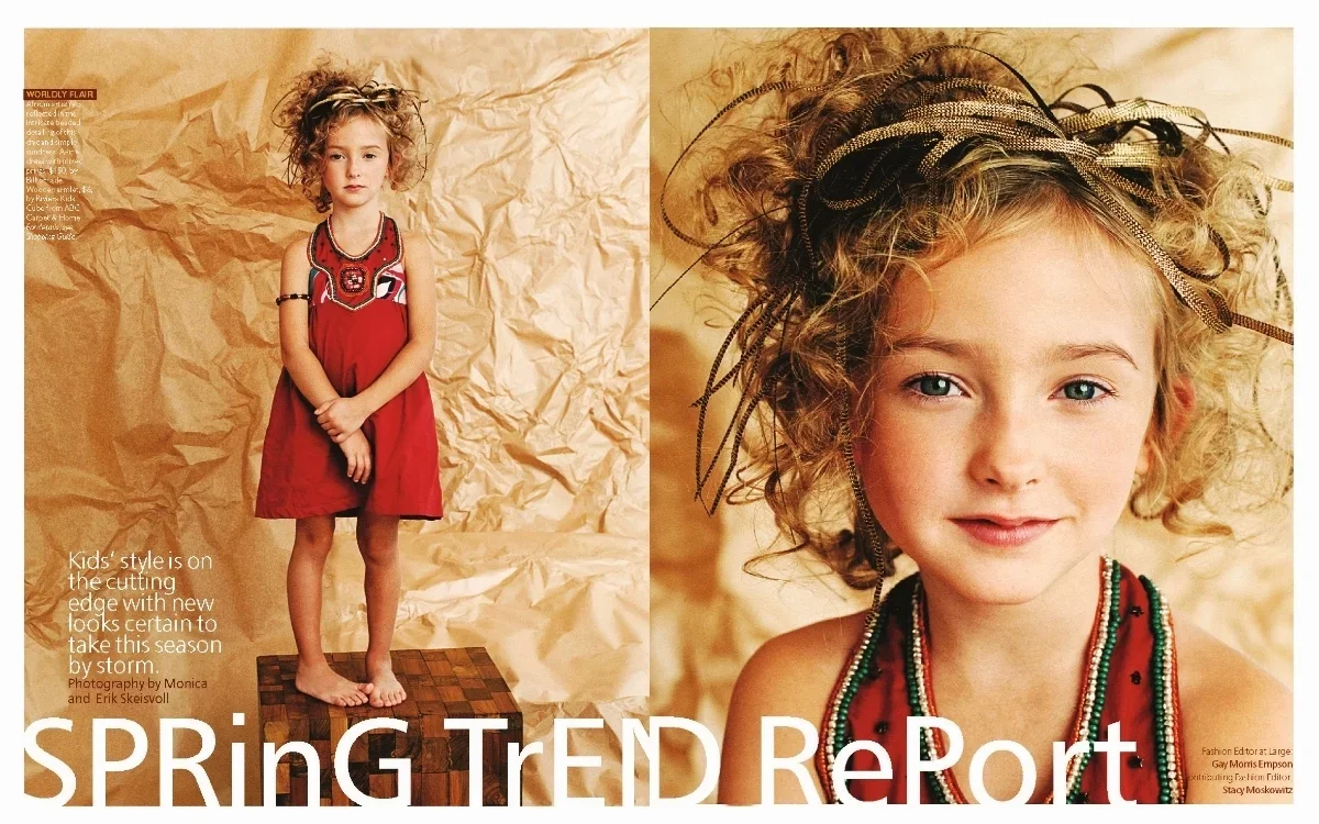 A young girl with curly blonde hair stands barefoot on a wooden platform, wearing a red dress with black and white embellishments. The background is crumpled brown paper. A close-up of her smiling face shows her wearing colorful bead necklaces and a 