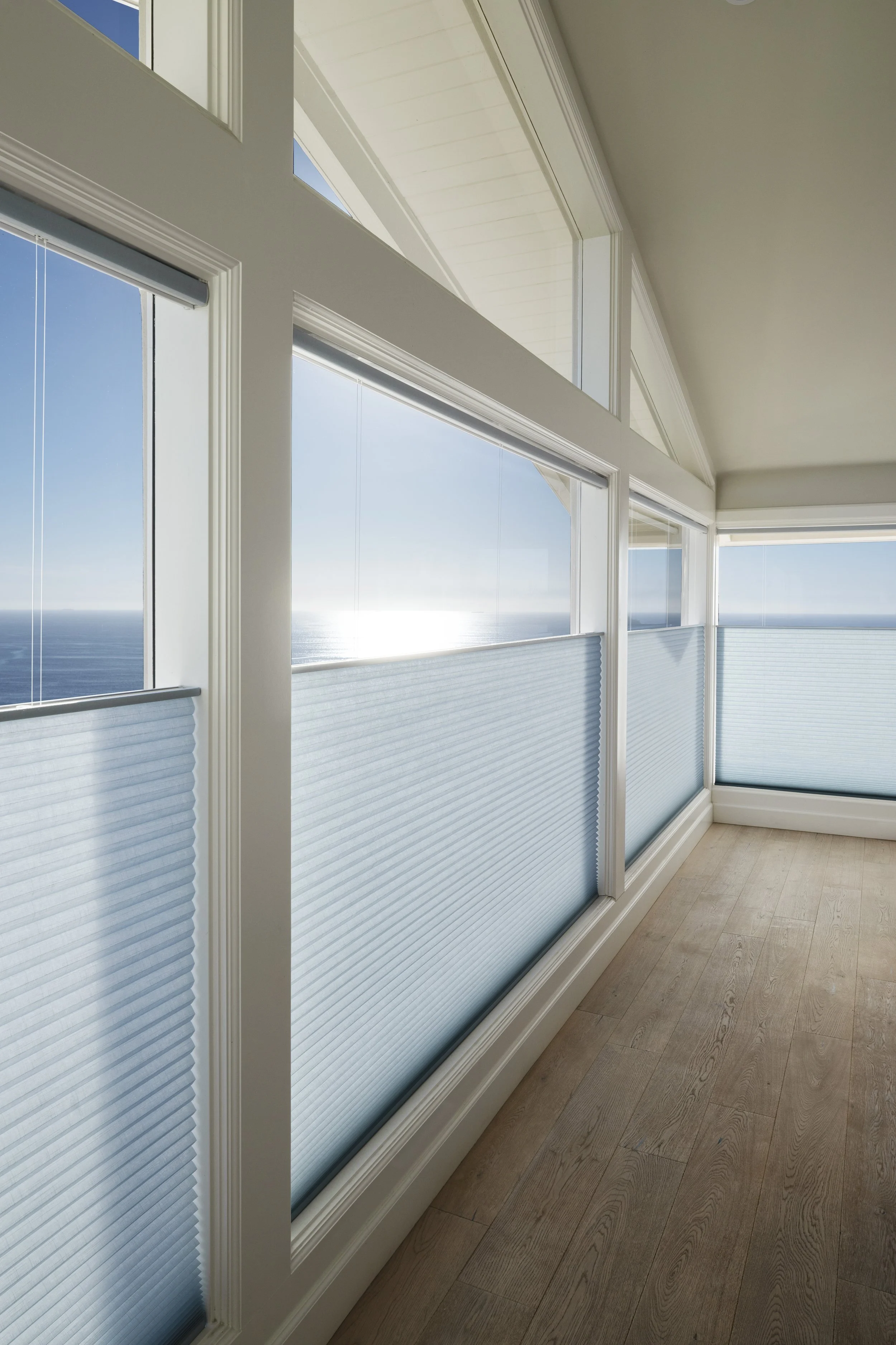 Interior view of a room with large windows showing an ocean view, with the lower parts of the windows covered by pleated blinds, and a wooden floor.