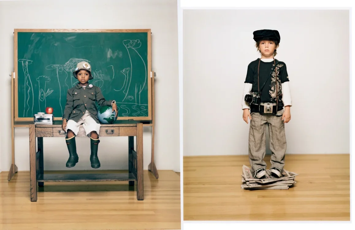 Two young children dressed as explorers or photographers: one sitting on a desk with a chalkboard behind, the other standing on a pile of newspapers with a camera around his neck, both wearing hats and rugged clothing.