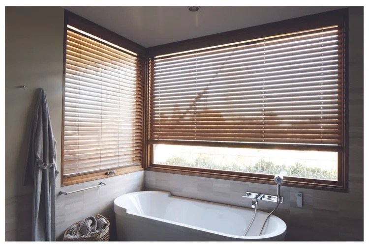 Bathroom with a white bathtub, wooden blinds on large windows, a towel hanging on a hook, and a basket with towels.
