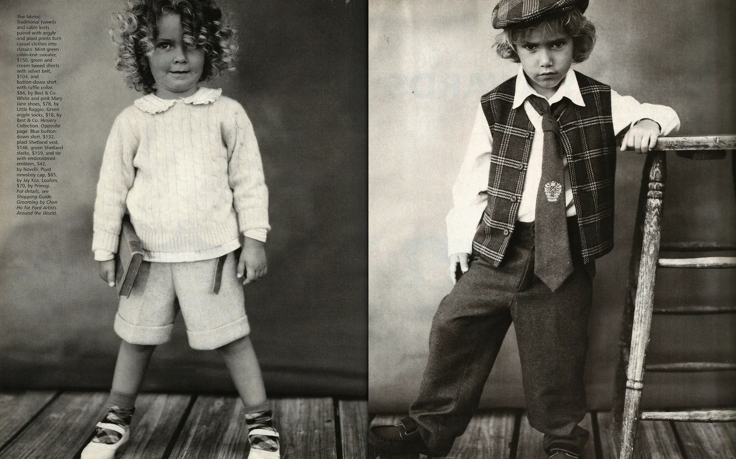 Black and white photo of two young children dressed in vintage clothing, standing against a plain backdrop. The girl on the left has curly hair and is wearing a sweater, shorts, and sandals, holding a book. The boy on the right has wavy hair, a cap, 