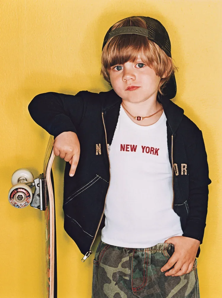 A young boy with shoulder-length hair, wearing a black snapback cap, a black jacket with jewelry, a white t-shirt with red text that says 'NEW YORK,' and camouflage pants, standing against a yellow background, holding a skateboard.