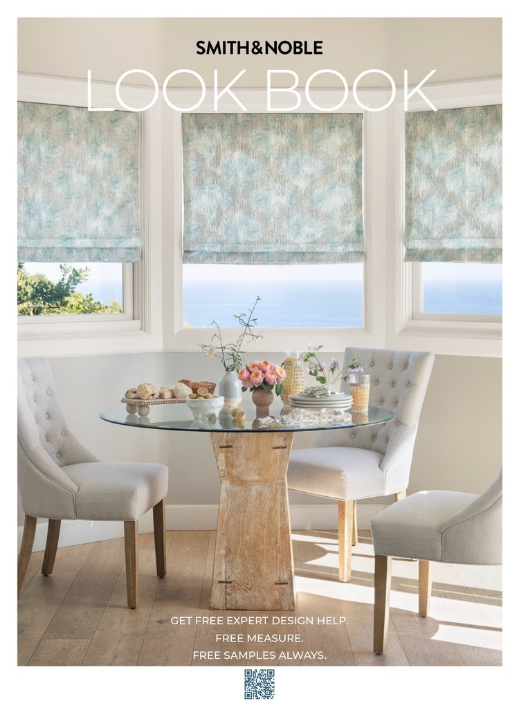Cover of a look book for Smith & Noble featuring a bright dining area with a round glass table, three upholstered chairs, three large windows with blue patterned shades, and ocean view in the background. The table has vases with flowers, stacked dish