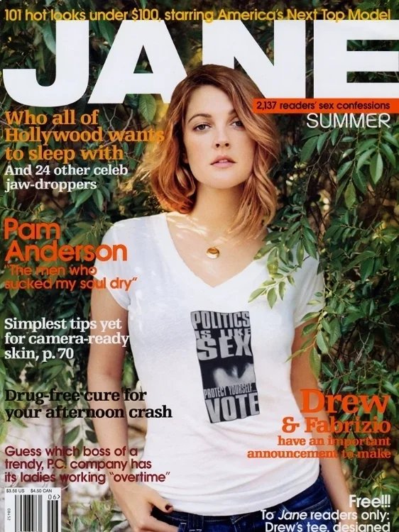 Woman with shoulder-length red hair standing outdoors in front of green foliage, wearing a white T-shirt with a political message and a chain necklace, looking at the camera.