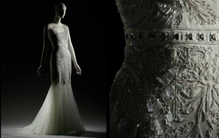 A mannequin wearing an elegant white wedding gown with intricate beadwork and embroidery, displayed against a dark background with a close-up of the detailed fabric.