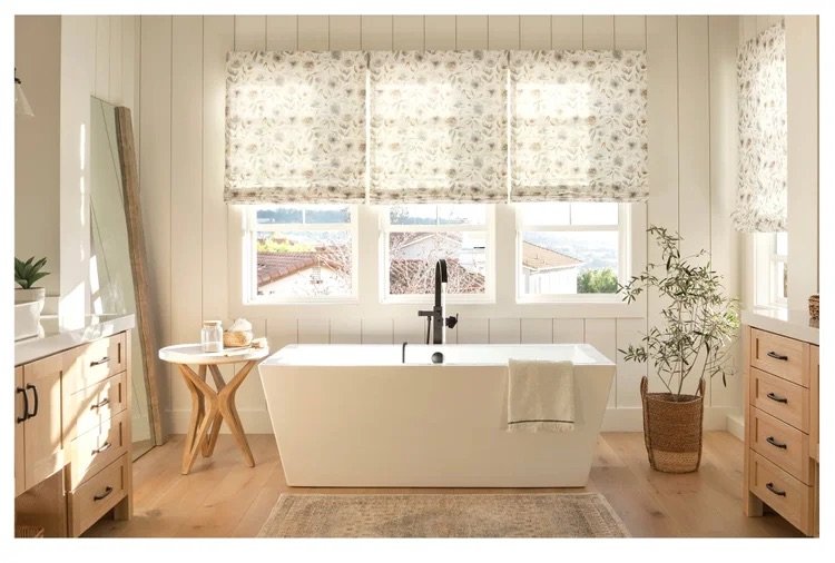 Bright bathroom with a central white bathtub, a towel hanging over the side, wooden cabinets, a small side table, a large mirror, potted plant, and patterned window blinds.