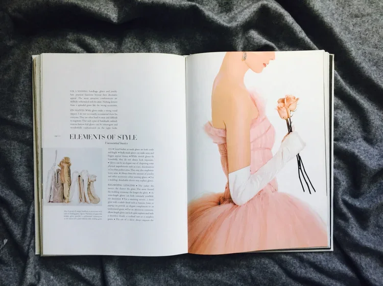 Open magazine on gray fabric, featuring a woman in a pink tulle dress holding a flower with long white gloves, and a page with text and images about fashion style.