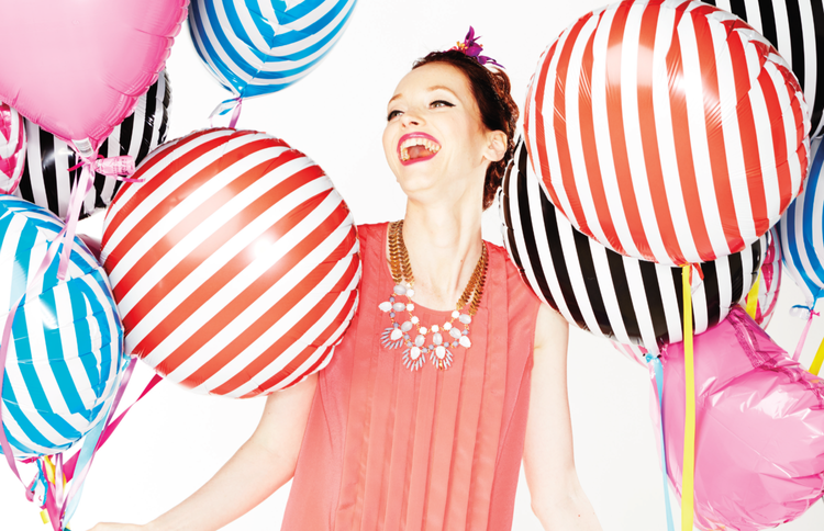 A woman smiling joyfully surrounded by colorful striped and pink balloons at a celebration or party.