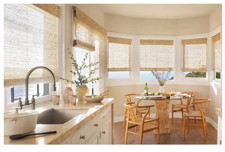 Bright dining area with round table, six wooden chairs, and large windows with woven blinds, sunlight streaming in, and a kitchen counter with a sink, plants, and decor.