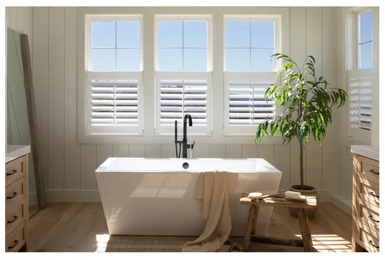 A bright bathroom with a white bathtub under three large windows with white plantation shutters. A towel hangs over the side of the tub, and a small wooden table with soap and towels is next to a potted plant.