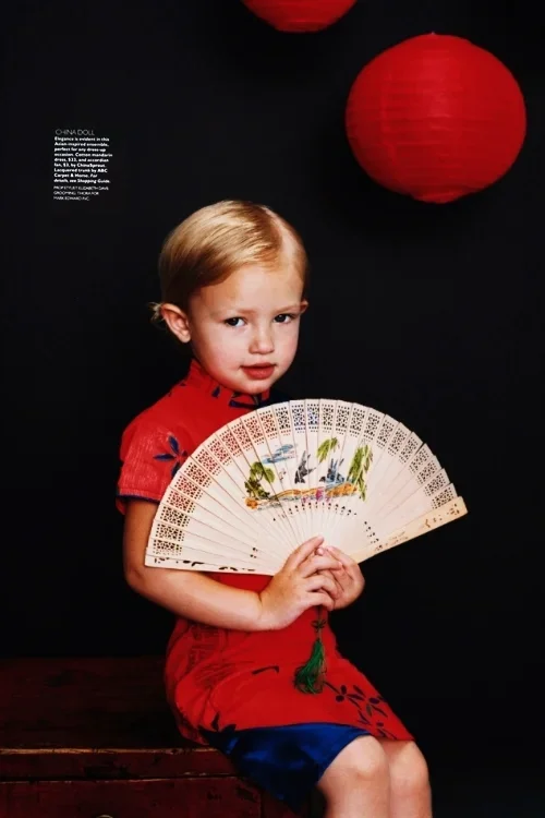 A young girl in a red dress with black and green details sitting on a wooden surface, holding a decorated folding fan, with a black background and a large red paper lantern hanging above her.