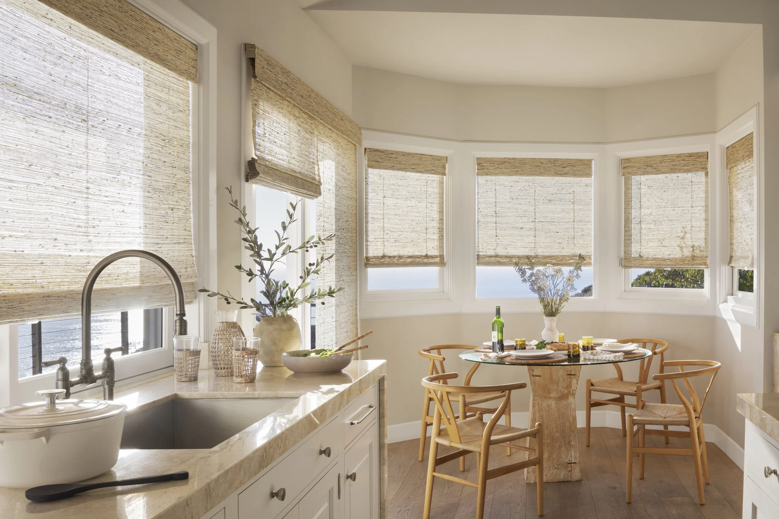 A bright kitchen and dining area with multiple windows covered by woven blinds, a white countertop with a sink, potted plants, a bowl, a green bottle, and a dining table set with plates, napkins, and glasses surrounded by wooden chairs.