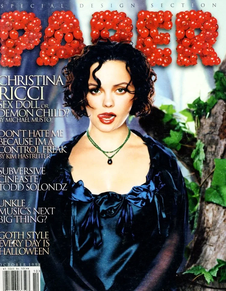 Magazine cover featuring Christina Ricci with short curly black hair, wearing a dark blue satin dress and green jewelry, with a background of green foliage and the word 'PAPER' made of red berries at the top.