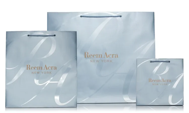 Three shopping bags with Reem Acra New York logo, varying in size, light blue color with silver handles.