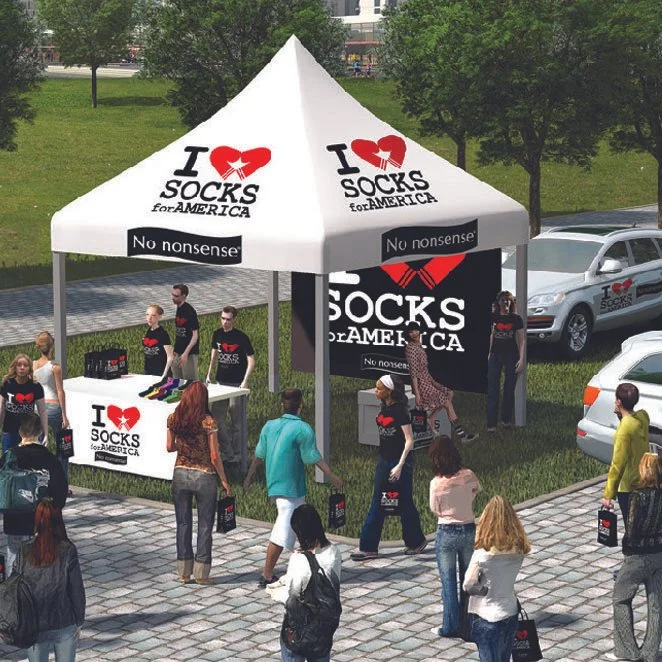A white tent with black and red 'I ♥ Socks for America' branding at an outdoor event, with people shopping and standing around, holding shopping bags, and cars parked nearby.