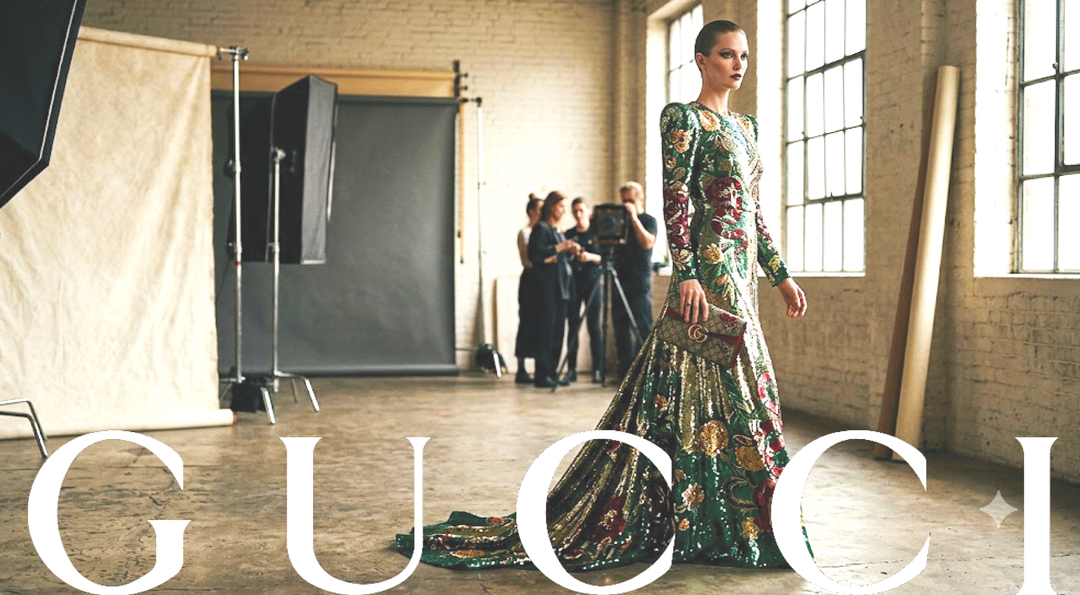 A woman in a colorful, ornate gown walking on a runway with a Gucci clutch bag, in a studio setting with large windows and a photography crew in the background.