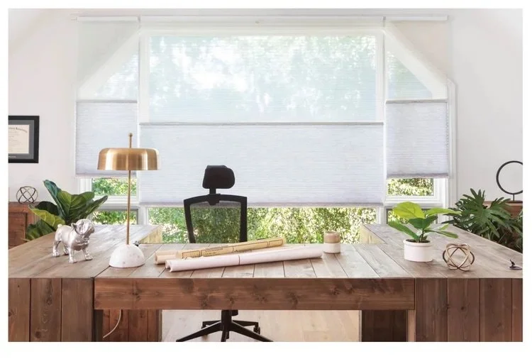 Home office with a wooden desk, black office chair, and window blinds with greenery outside.