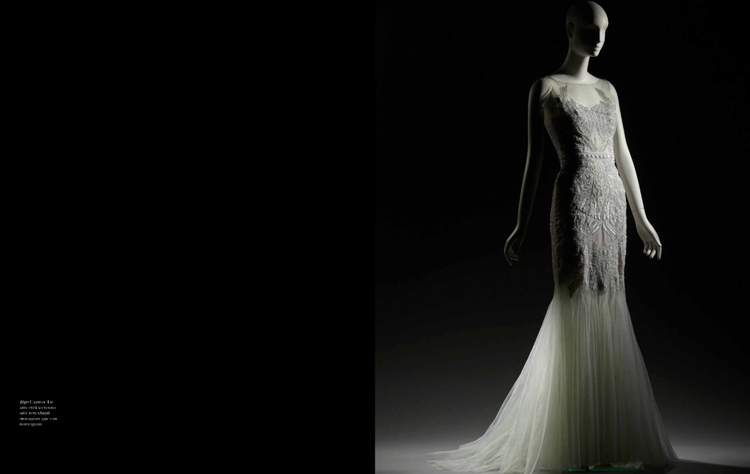 A mannequin wearing an elegant white wedding dress with lace details and a long flowing train, illuminated against a dark background.