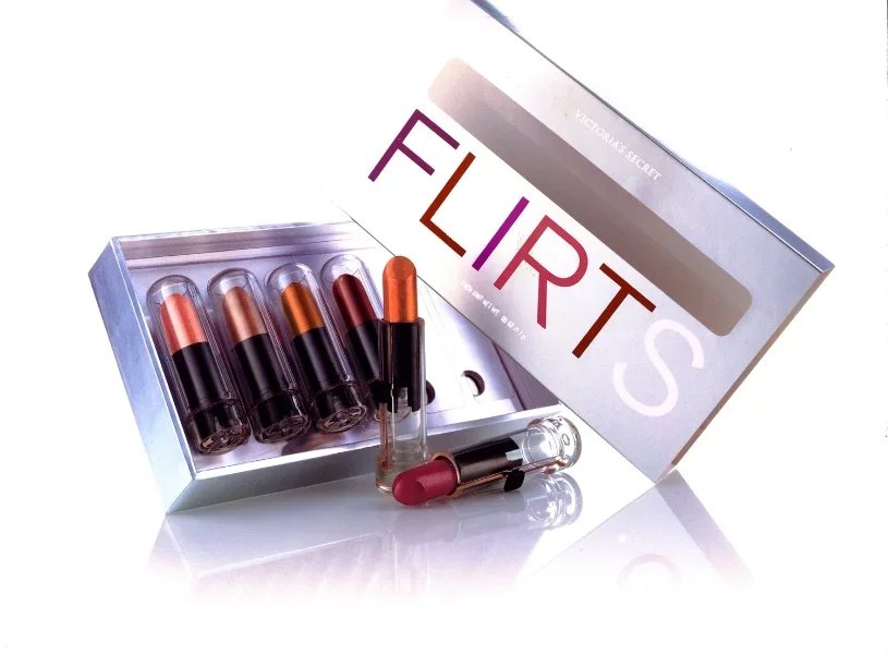 Open box of Victoria's Secret FLIRTS lipsticks in various shades with one lipstick taken out and lying on the surface.