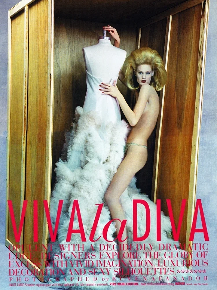 A minimalistic fashion magazine cover featuring a woman with voluminous blonde hair, minimal clothing, and bold makeup, standing next to a mannequin with a white dress and fluffy white fabric. The background is a wooden box-like frame, and large pink