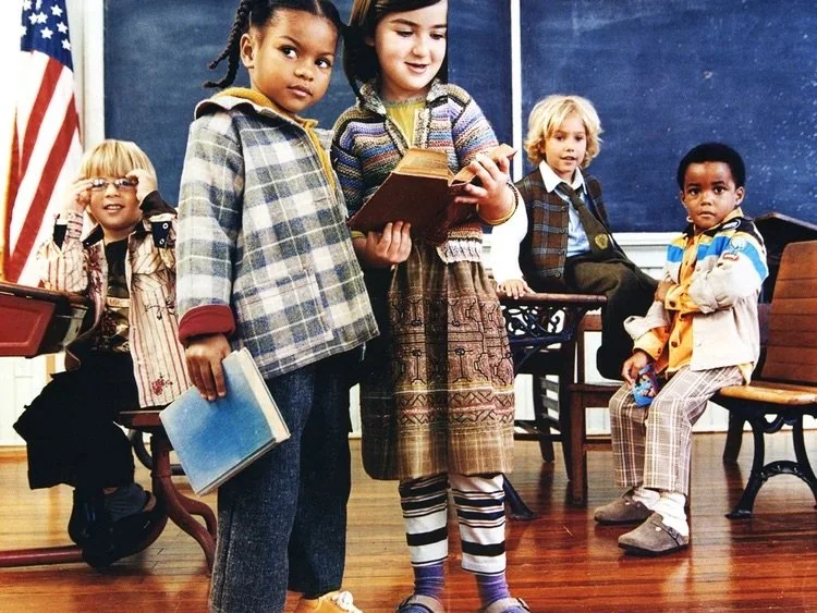Five children in a classroom, some standing and some sitting, with an American flag on the left and a chalkboard in the background. Two girls are in the foreground, one holding a book and the other with a book in her hand. Three boys are behind them, one sitting at a desk with a toy and headphones, another sitting on a bench, and the third watching.