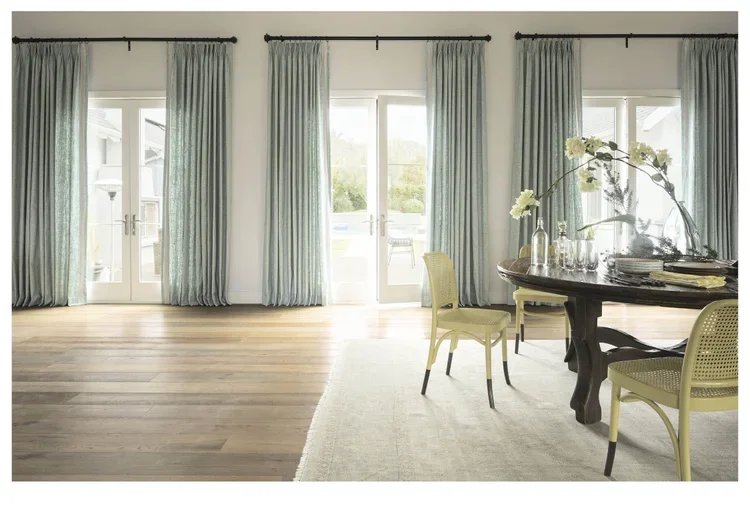 Bright living room with light blue curtains, large glass doors, and a round dining table with yellow chairs and decorative flowers.