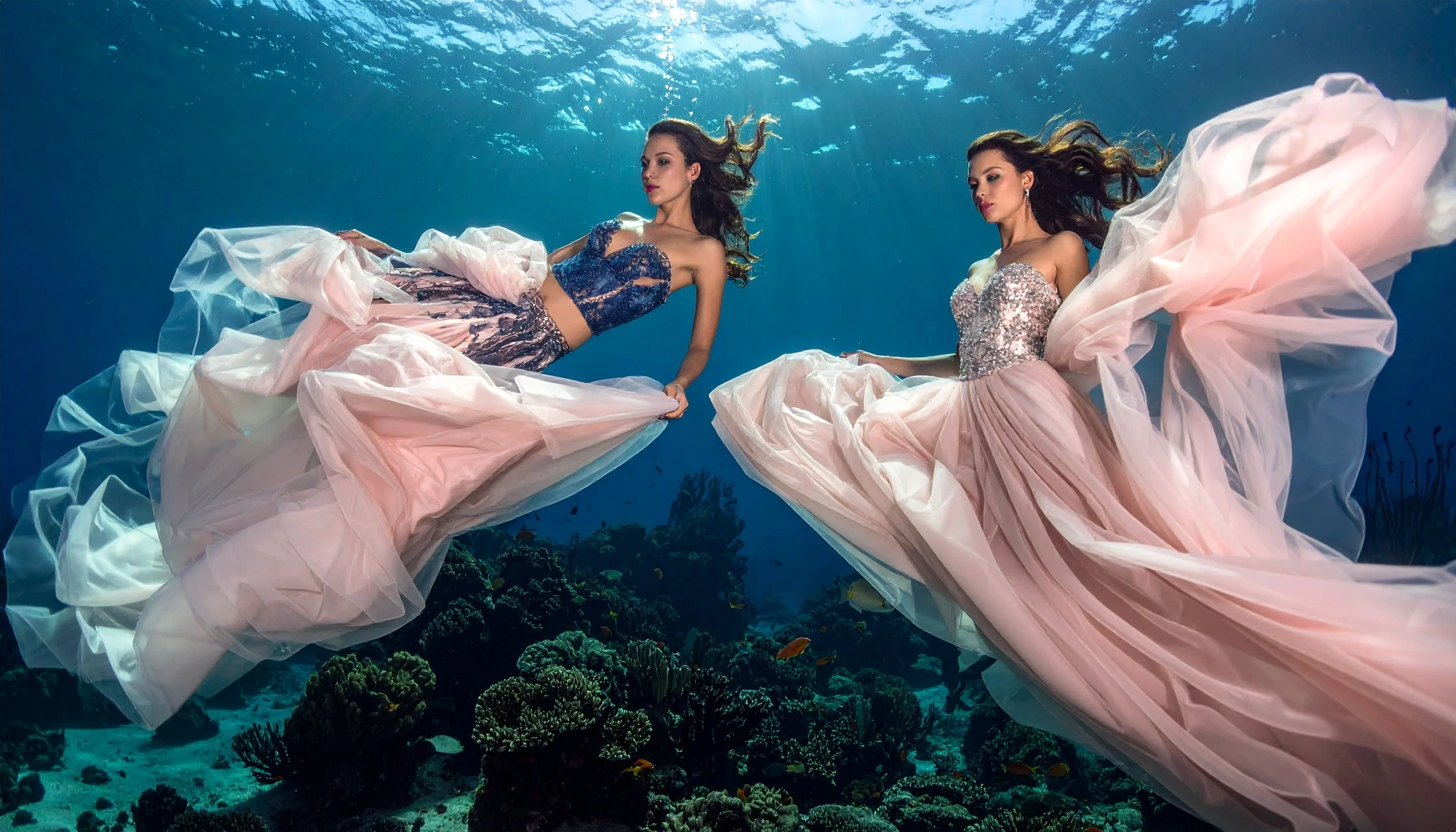 Two women in elegant dresses swimming underwater in a coral reef.