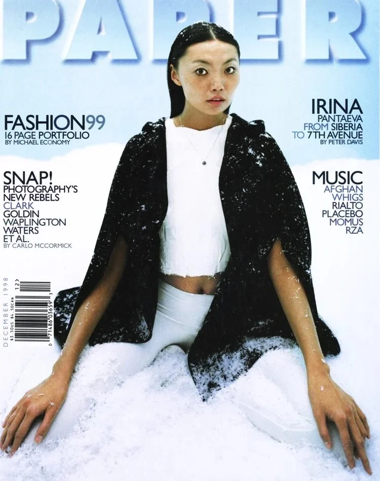 Young woman with wet hair and a surprised expression, sitting in snow, wearing a white crop top, white pants, and a black oversized jacket, on the cover of Paper magazine.