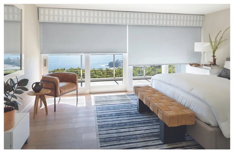 Modern bedroom with large windows showing a view of water and greenery, a bed with white bedding, a striped rug, a small side table with a plant, a wooden bench at the foot of the bed, and a comfortable armchair near the window.