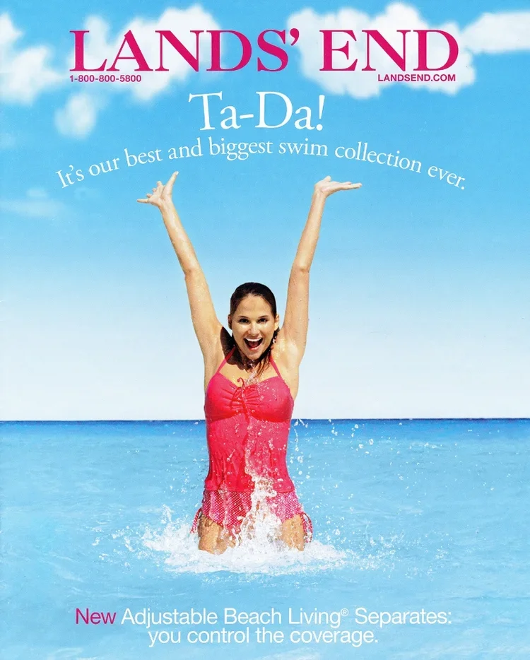 A woman standing in the ocean water with her arms raised, smiling, wearing a pink swimsuit against a blue sky with clouds. The image is an advertisement for Lands' End, promoting their swim collection and adjustable beach living separates.