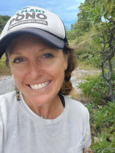 Woman wearing a cap that says 'Plant Pono' and a white t-shirt, smiling outdoors next to green leafy plants or trees.