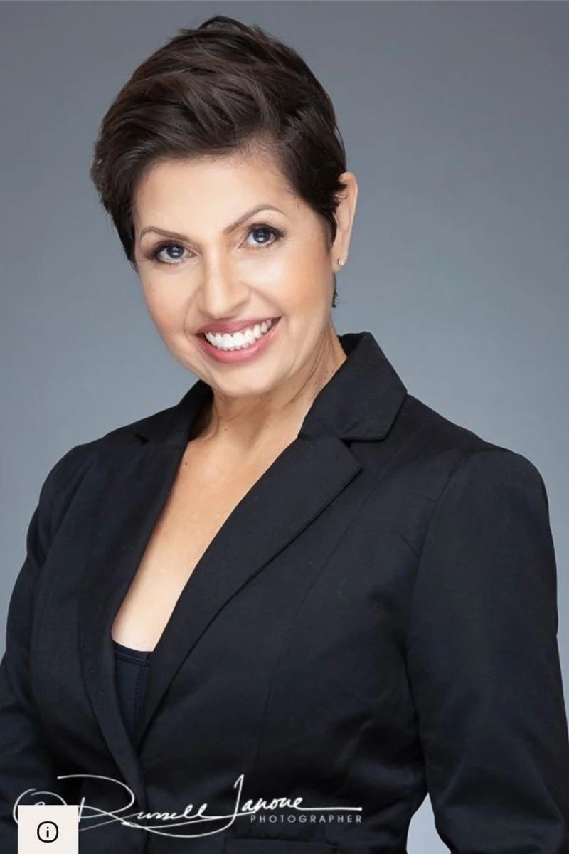 A woman with short dark hair smiling, wearing a black blazer against a gray background.