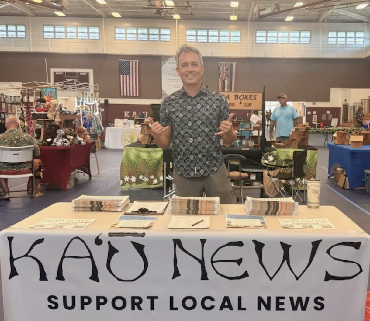 A man standing behind a table with a banner that reads 'KAU NEWS SUPPORT LOCAL NEWS' at a market or fair. The table has stacks of newspaper and pamphlets. In the background, people browse vendor booths with various items, and flags are hanging on the walls.