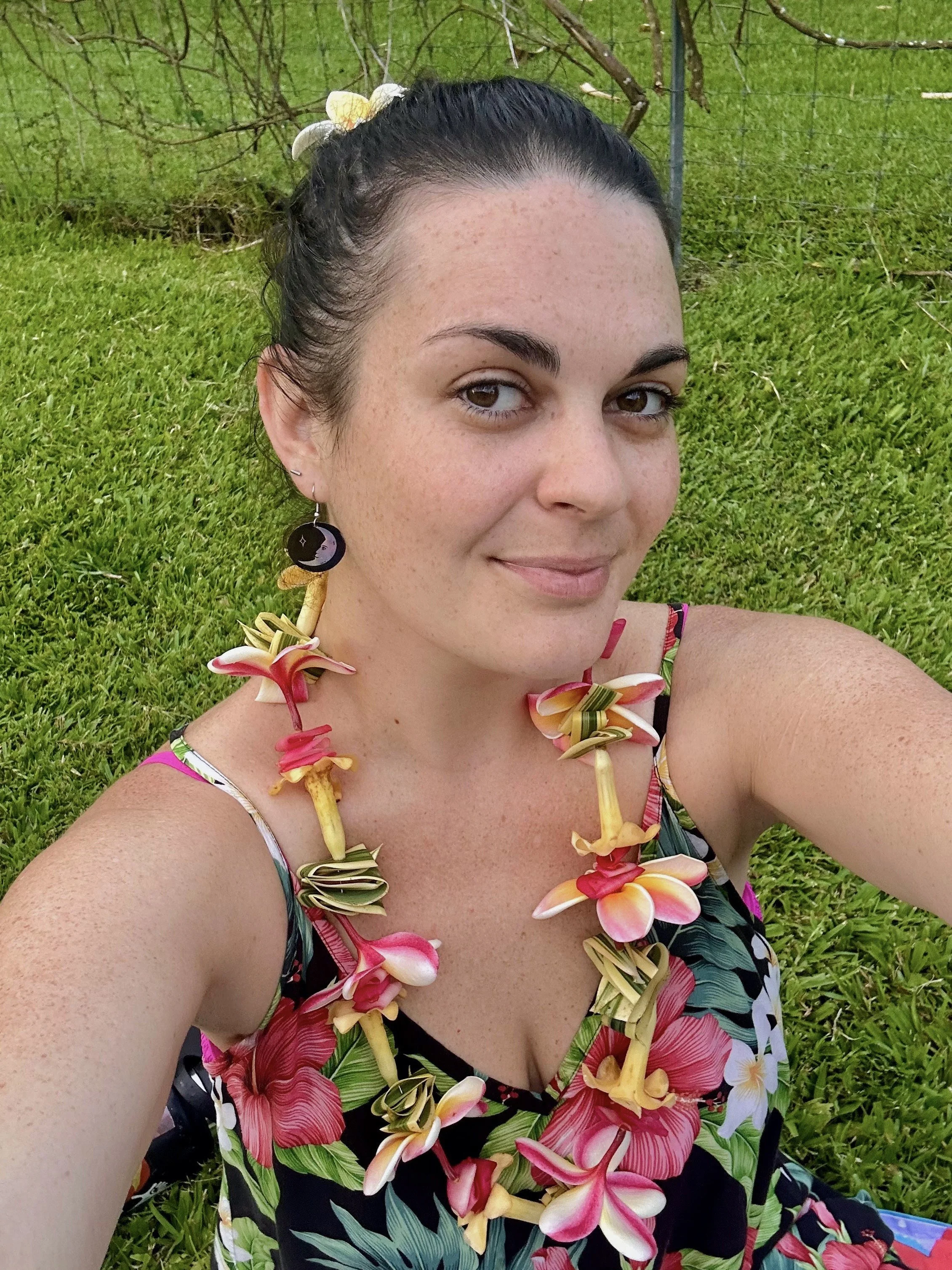 A woman wearing a floral dress and a lei made of pink and yellow flowers, with a black earring featuring a moon and star design, taking a selfie outdoors on a grassy area with a fence and greenery in the background.