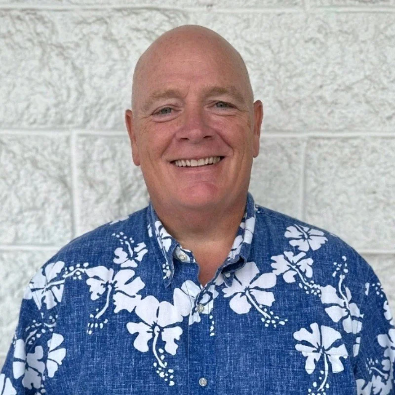A smiling man wearing a blue and white Hawaiian shirt, standing in front of a light-colored brick wall.