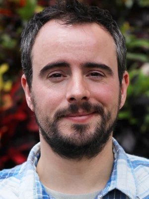 A man with short dark hair and a beard, smiling slightly, wearing a light blue and white checkered shirt outdoors with blurred green and red foliage in the background.