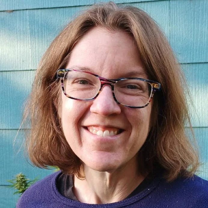 A woman with brown hair, glasses, and a purple sweater standing in front of a blue wooden wall, smiling at the camera.