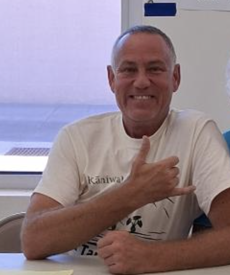 Man smiling and making a shaka hand gesture while sitting at a table in a room with windows.