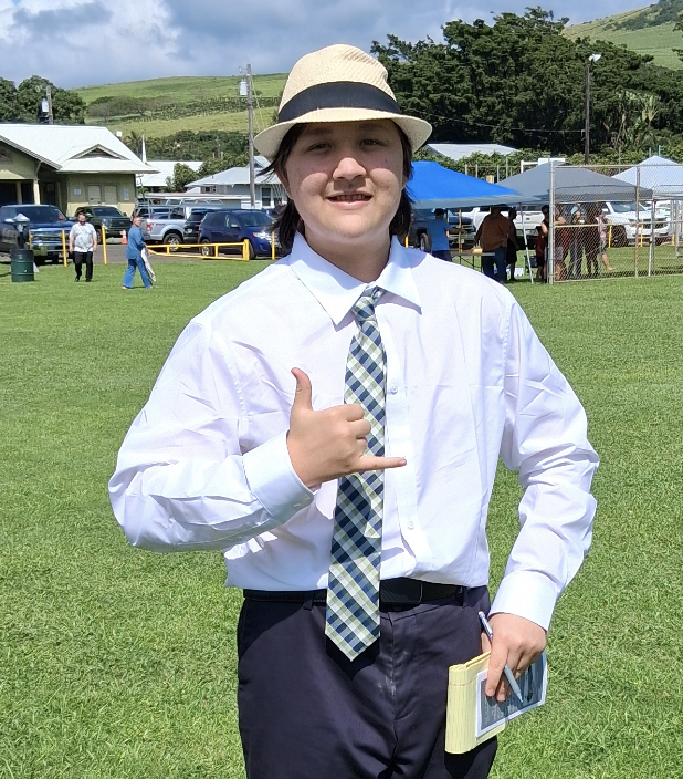 A young person in a white dress shirt, striped tie, black pants, and a tan fedora hat, standing on a grassy field with outdoor tents and people in the background, making a shaka hand gesture and holding a notebook and smartphone.